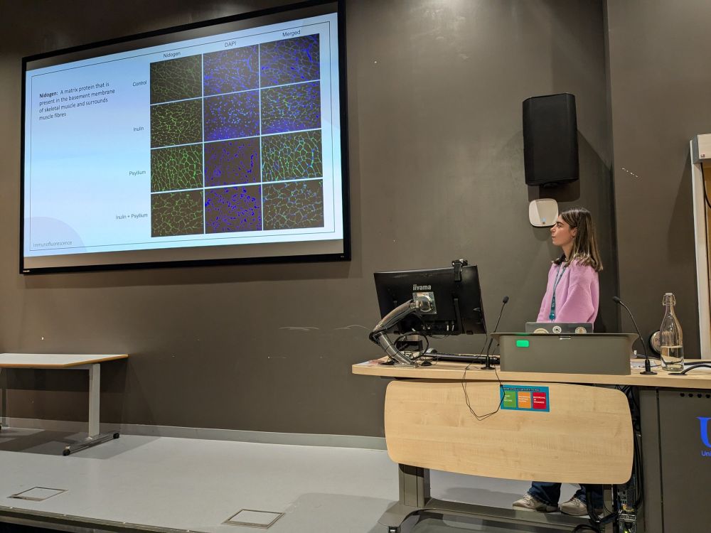 Helena at a podium in front of a large screen showing an image of her results from immunocytochemistry analysis of mouse muscle in response to different levels of fibre in the diet.