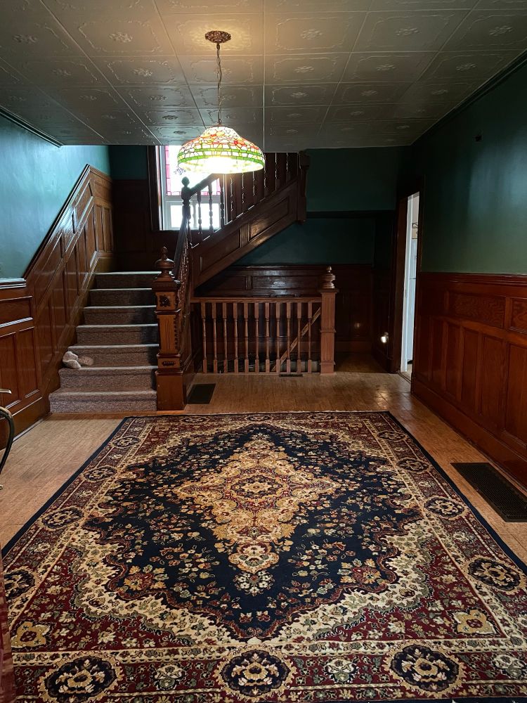 A victorian era room with wood wall panelling, a staircase with wooden railings, bluish green walls, a tiffany pendant lamp and ceiling tiles painted a very pale mint. There is a persian rug on the floor.