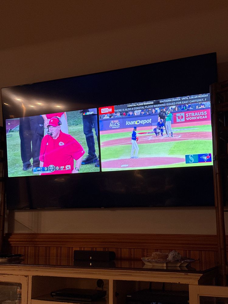 A split screen TV showing the Chiefs/Lions game on the left and the Blue Jays/Mariners in the ALCS on the right. The football game shows Andy Reid on his headphones. The baseball has the Mariners at bat and Gausman pitching for the Jays.