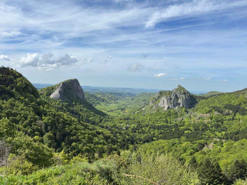 Paysage de montagne avec deux formations rocheuses entourées de forêts verdoyantes sous un ciel bleu légèrement nuageux. La vallée verte entre les montagnes offre une vue panoramique impressionnante.
