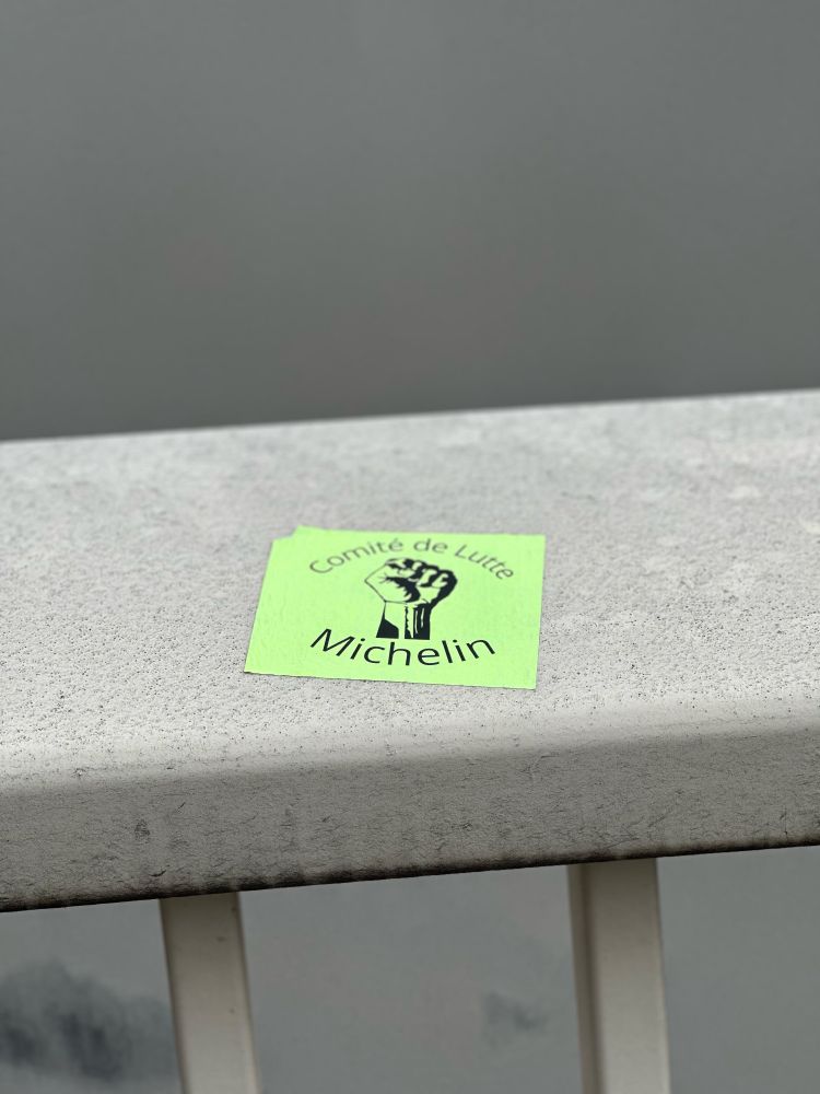 Cette image montre un autocollant vert collé sur une rambarde grise. Sur l'autocollant, il y a un graphique noir représentant un poing levé accompagné du texte « Comité de Lutte Michelin ».