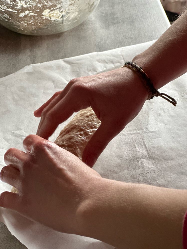 Deux mains en train de façonner une pâte à pain sur une feuille de papier cuisson. 