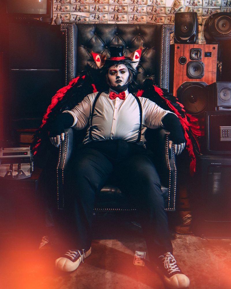 A photograph of a Husk cosplay from the Amazon Prime Series Hazbin Hotel, with Husk reclining in a throne like chair surrounded by old televisions and audio equipment.