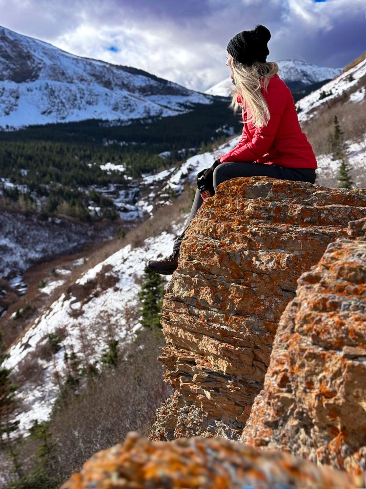 A woman in winter clothes sits on a mountainside, with snowy mountains below her. 