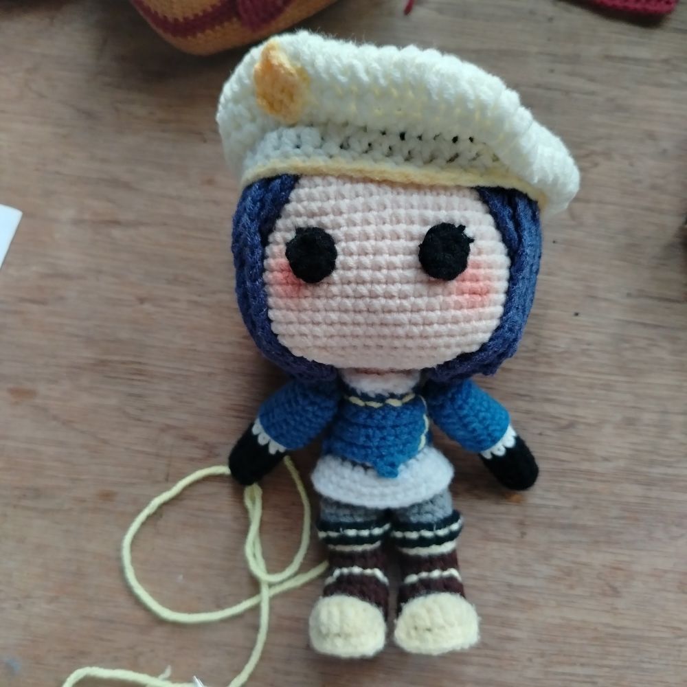 Crochet League of Legends character, commander caitlyn, laying on a wodden desk. The doll has black eyes and gloves, blue hair and coat, white hat and skirt, brown shoes, and yellow accents. It is still missing some detailing and her cape