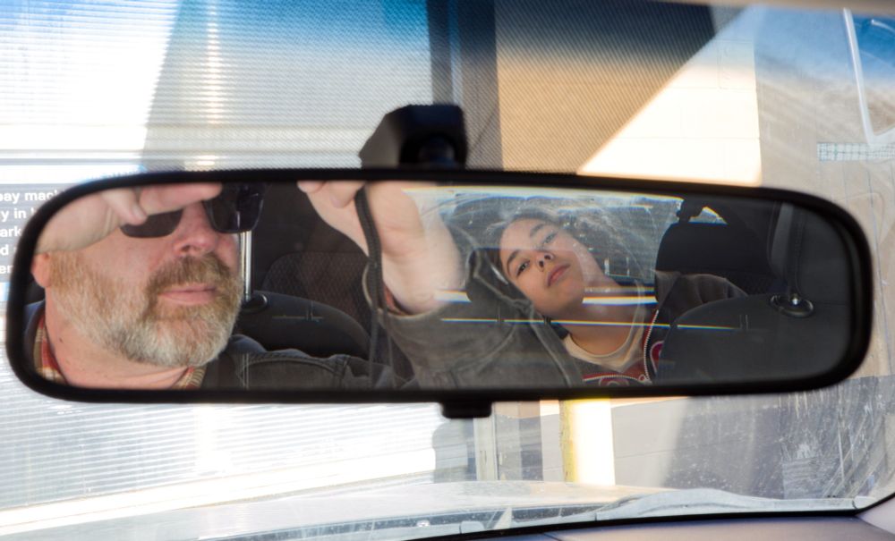 A father takes a photo of their son in the review mirror of a vehicle. 
