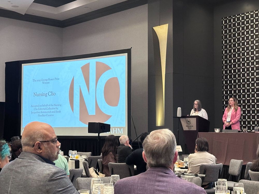 A conference room scene showing an awards ceremony. On stage, two women stand at a podium, one speaking and one standing to the side. A large screen behind them displays a slide that reads: “The 2025 George Rosen Prize Winner: Nursing Clio. Accepted on behalf of the Nursing Clio Editorial Collective by Jacqueline Antonovich and Sarah Handley-Cousins.”