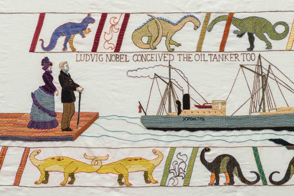 Upper and lower embroidered borders show a frieze of dinosaurs, purple, yellow, green, and black (there are two yellow dinosaurs, their tails intertwined in a kind of lovers' knot). In between we see a Victorian lady and gentleman standing on a pier, looking at a huge steam ship in the sea. The caption is Ludvig Nobel Conceived the Oil Tanker too.
