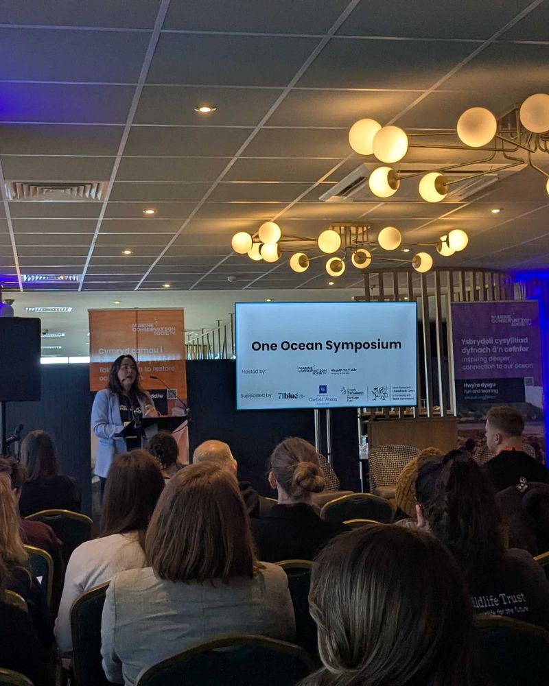 Photo taken at the One Ocean Symposium of a room filled with people sat down listening to a speaker stood at the front of the room.
