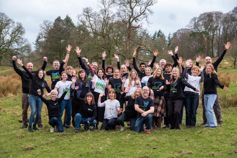 Rewilding Britain smiling outdoors, wearing Rewilding Britain shirts, standing on grassy terrain with trees in the background in the Lake District. Credit: Katherine Gulika.