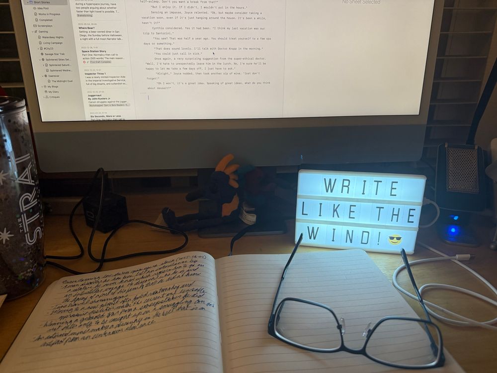 A photo of my writing desk, with a lit-up mini movie marquee with the message “WRITE LIKE THE WIND! 😎”. Also visible in a notebook with a pair of computer glasses resting on it, and the lower half of a computer screen, along with a pair of plush kokopeli figures sitting back to back on the desk.