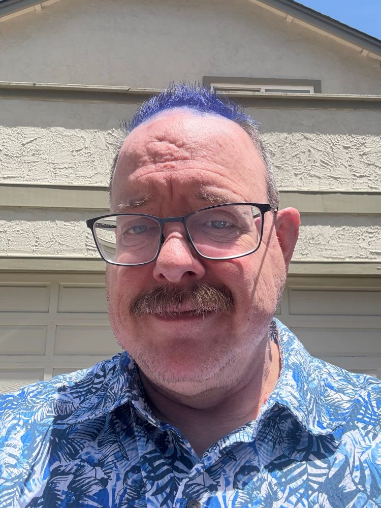 A selfie of me standing in the sun, wearing a brightly patterned blue and white shirt, with the hair on the top of my head dyed cobalt blue. I’m /almost/ smiling.