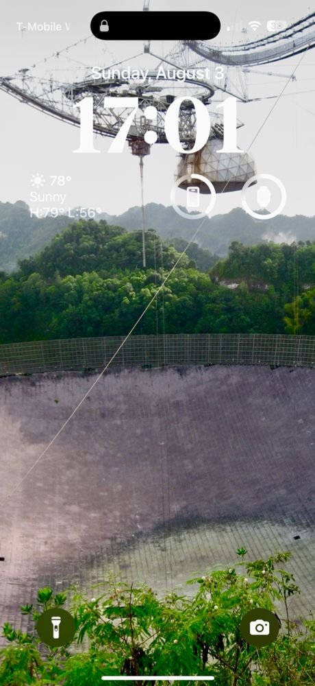 A screenshot of my lockscreen taken at 17:01, with a vacation photo of a large radar dish built into a small valley surrounded by jungle, with the transceiver module hanging above it.