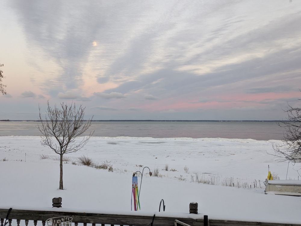 The scene is snow in the foreground and then some shore ice building up and then the Waters of Northwest Lake Michigan and the Stonington Peninsula is in the background and above them are clouds that have shades of pink from the setting Sun and the full moon is rising above it all and partially shrouded by clouds