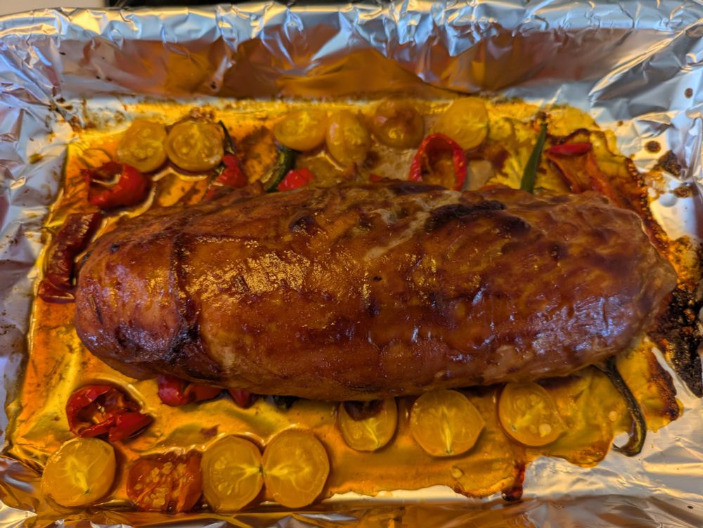 A roasted pork tenderloin shown in a pan that's lined with aluminum foil and the roast is marinated and deep Brown and was roasted on a bed of spicy hot peppers. Sun gold, yellow cherry tomatoes are also in the pan which provide some sweetness to go with the heat from the hot peppers. 