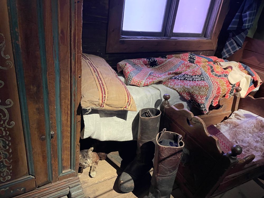 Interiör från en historisk bondstuga. Säng, skåp, stövlar och en vagga. Skåpet och sängens täcke har dekorativa mönster och färger.