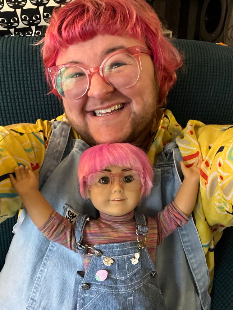 a selfie of emmett, a trans man with pink hair, a scruffy beard, and pink glasses. he is smiling. in his lap is an american girl doll who has been customized to look like him. emmett and his doll are dressed in matching colorful shirts and denim overalls. 