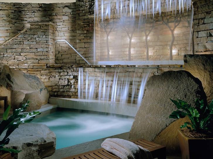 A plunge pool at a spa, set between 2 large rocks and a waterfall cascading down. Soft lighting helps with the relaxing mood. 