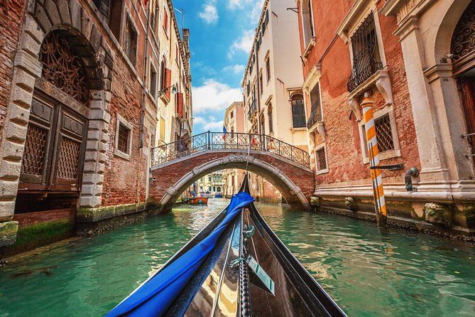 A picturesque view from a gondola, between tall historical buildings in Venice. A cute little bridge across the water stands just in front of the boat. A blue sky peeks out just between the buildings and the golden light from the sun shines through. 