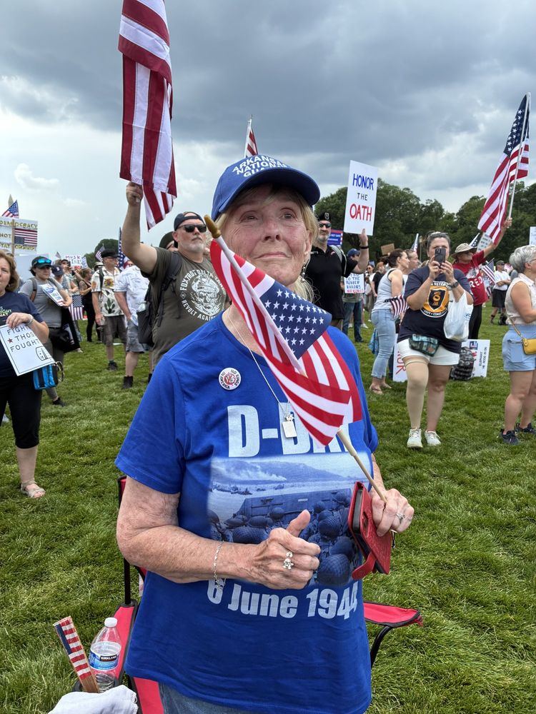 A woman wearing a D-Day t-shirt and an Arkansas National Guard ballcap is holding an American flag. In the background, there are others with signs and flags.