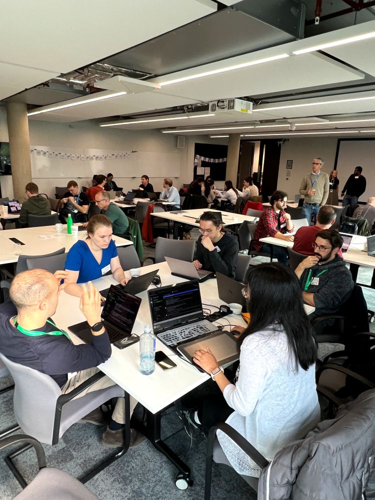 In a busy meeting room, participants are seated at white tables working on laptops, working independently or in groups. 