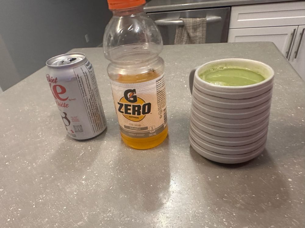 Three open drinks sit upon a grey counter in a neat row. To the left, a Diet Coke in a can. In the middle, an orange Gatorade (zero sugar) half consumed. On the right, a grey mug full of matcha tea. 