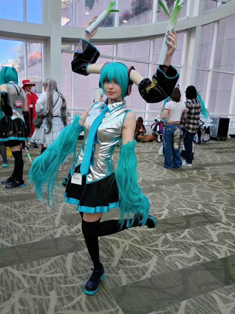 A Hatsune Miku cosplayer who has put the wig ponytails on her arms and has sculpted arms holding leeks on her wig.