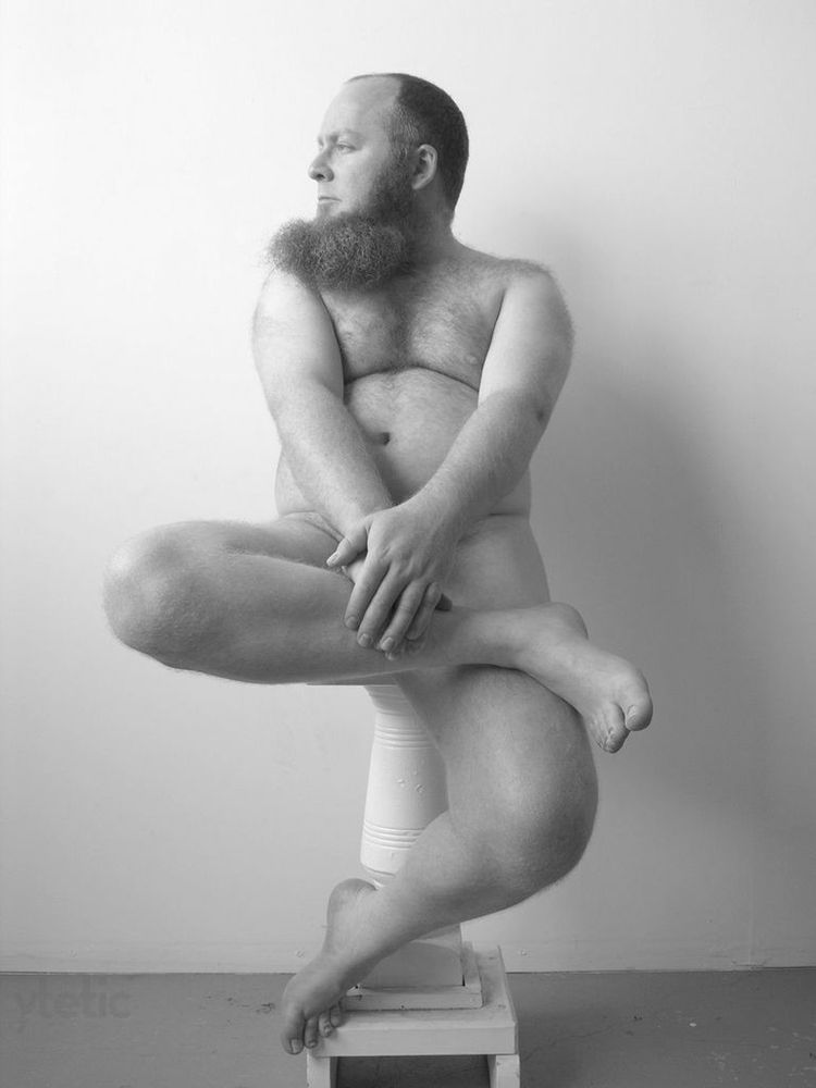 BW photo, studio. Sitting nude on the posing stand, one foot touching the lower part of the pedestal, the other drawm up and held by both his hands He's looking out of frame to the left.