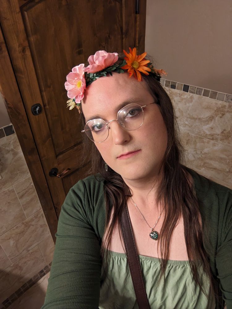 A woman takes a selfie in a green dress, shawl, and flower crown.