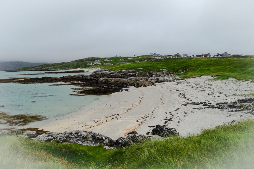 Hiekkaranta, vaaleaa hiekkaa ja rantakallioita. Vasemmalla näkyy pala vaalean turkoosia merta. Edustalla ja rannan toisella puolen vihreää ruohikkoa. Kauempana hautausmaa ja kylän rakennuksia. Pilvinen taivas.
Eriskay, Ulko-Hebridit.