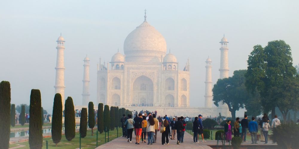 Ihmisiä kävelemässä kävelytietä pitkin kohti aamu-usvasta erottuvaa Taj Mahalia. Vasemmalla sypressipuiden takana toinen puiston kanavista, oikealla puolella puita.