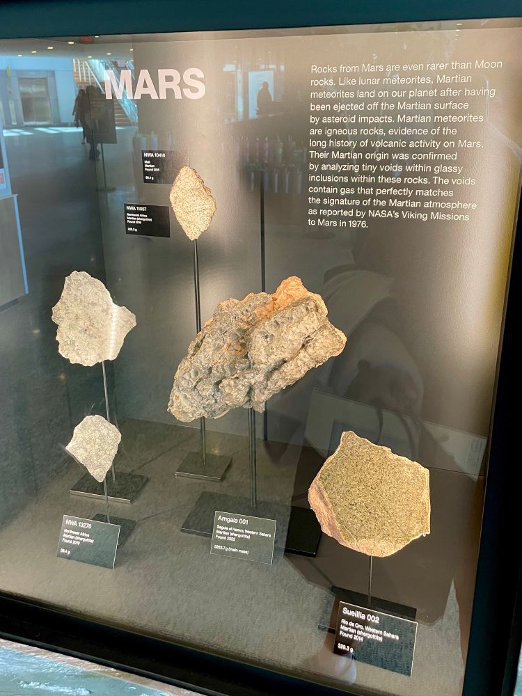 Exhibit with descriptor: “Rocks from Mars are even rarer than Moon rocks. Like lunar meteorites, Martian meteorites land on our planet after having been ejected off the Martian surface by asteroid impacts. Martian meteorites are igneous rocks, evidence of the long history of volcanic activity on Mars Their Martian origin was confirmed by analyzing tiny voids within glassy inclusions within these rocks. The voids contain gas that perfectly matches the signature of the Martian atmosphere as reported by NASA's Viking Missions to Mars in 1976.”