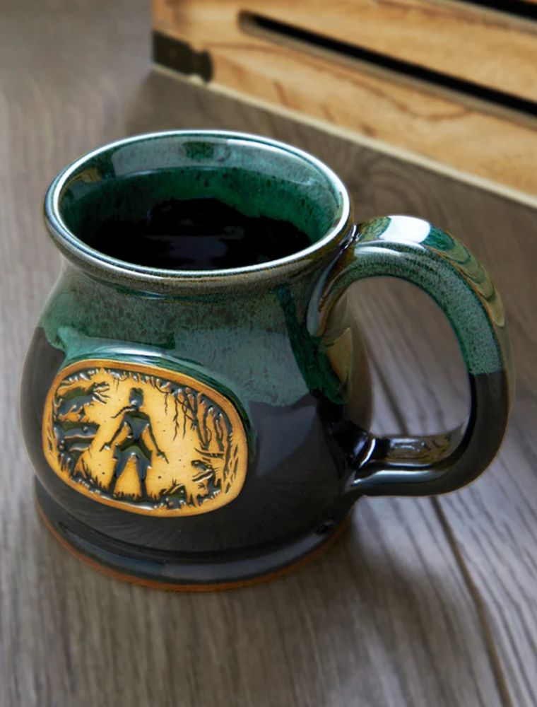 A royal green ceramic mug, with a yellow emblem in the center displaying Lara Croft in silhouette, surrounded by enemies. The mug has an elegant, curved shape and a glossy glaze on top. 