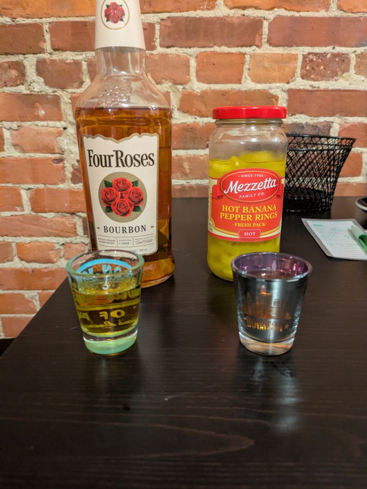 Bottle of four roses bourbon with a shot poured, next to a bottle of hot banana peppers and a shot of pepper brine.