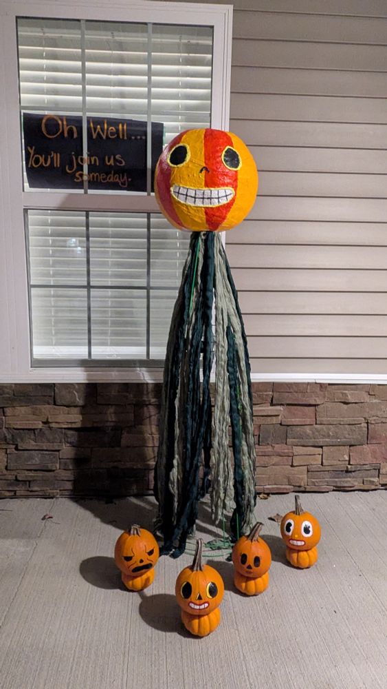 A giant pumpkin head with green streamers underneath surrounded by smaller painted pumpkin people. The sign in the window behind the characters reads, "Oh well....you'll join us someday." The characters are from the show Over the Garden Wall
