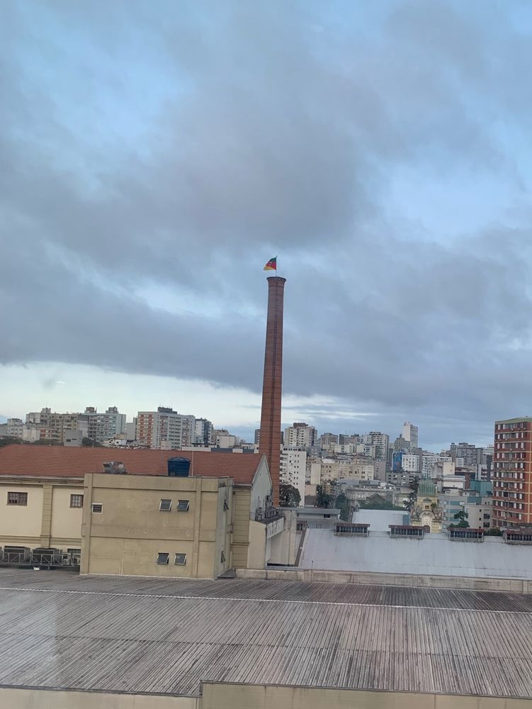 Vista do nono andar de um prédio, onde aparece o céu nublado, os telhados de outras edificações e uma chaminé com a bandeira do Rio Grande do Sul tremulando.