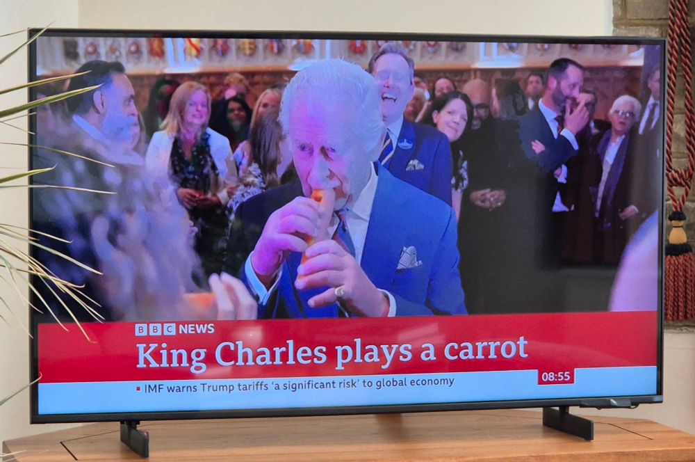 A TV showing BBC News, with a picture of King Charles playing a carrot in a similar manner to a recorder. The caption says "King Charles plays a carrot".