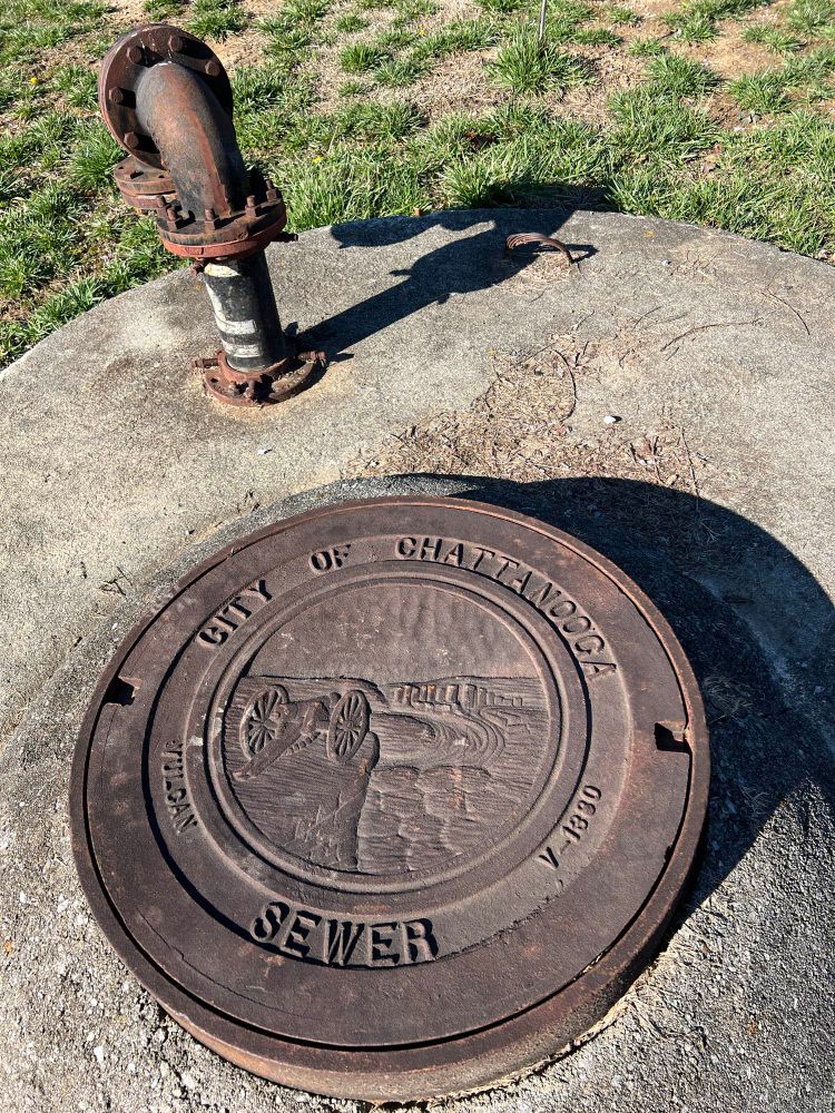 Sewer cover with “City of Chattanooga” and “SEWER” encircling a carving if a cannon on a ridge overlooking a river bend.  A pipe sticks ip out of the ground behind the sewer cover.