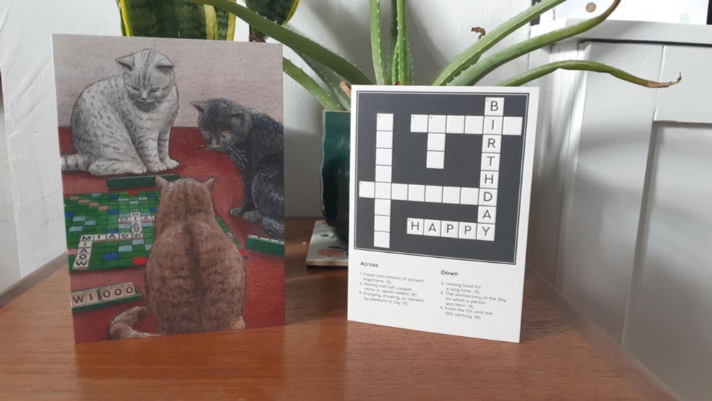 Two birthday cards, one showing cats playing Scrabble and one which is a crossword with HAPPY BIRTHDAY filled in