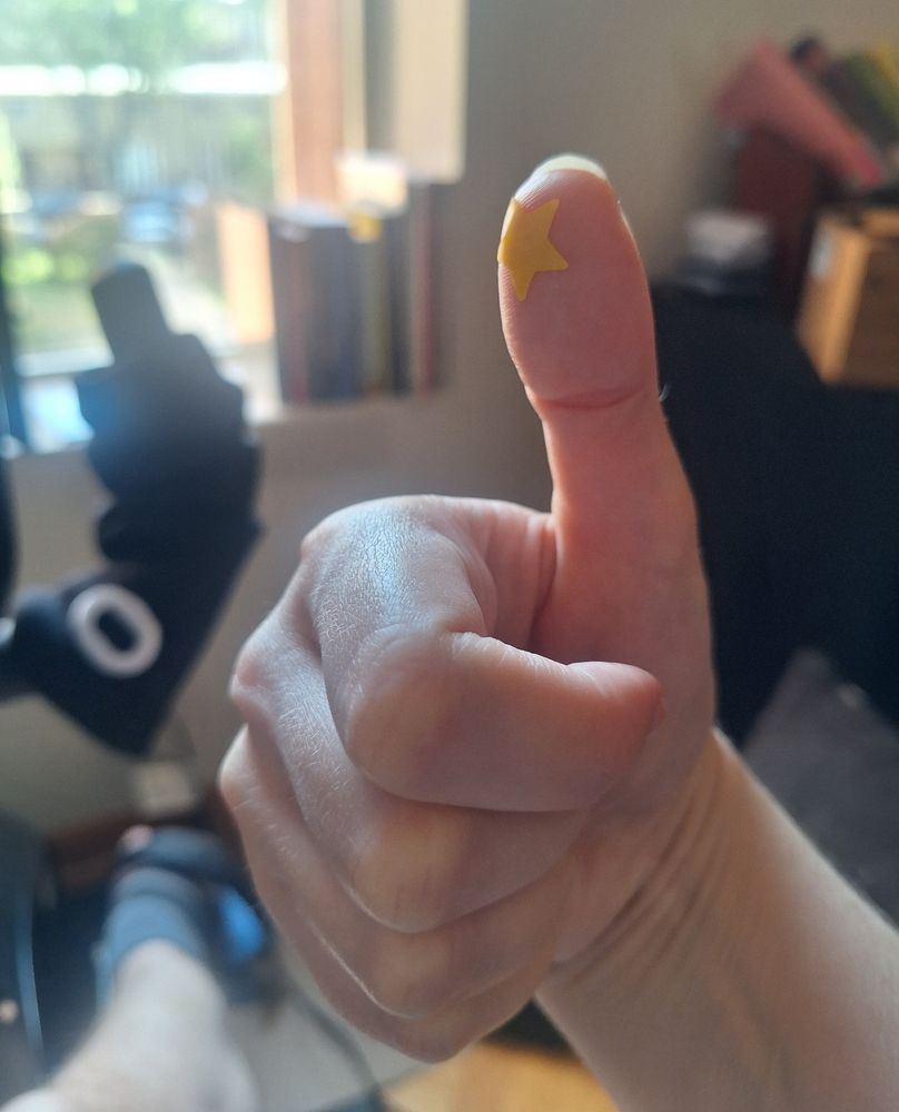 close-up of a pale hand giving a thumbs up against a blurred domestic background. The thumb's pad has a yellow star-shaped bandage on it 