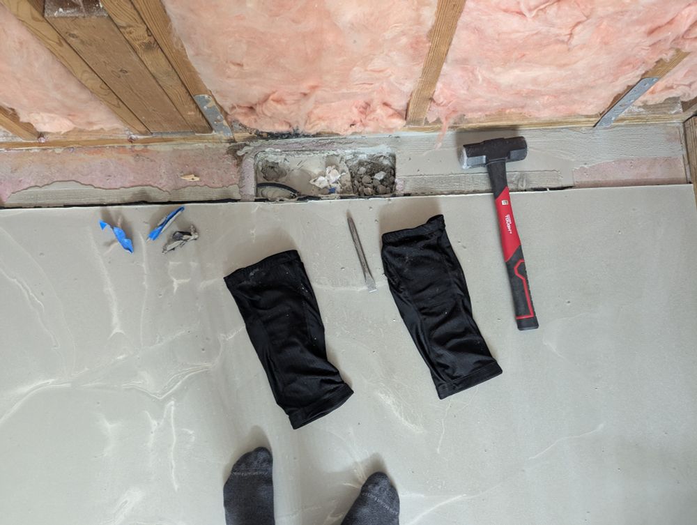 A photo of a bathroom floor with freshly poured self-leveling concrete. In the corner, the shower drain is barely visible, partially filled with toilet paper. The concrete has seeped underneath the makeshift barrier, covering part of the drain area that was supposed to remain open.