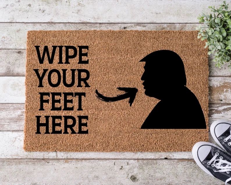 Coir doormat that says WIPE YOUR FEET HERE with an arrow pointing to a silhouette of Trump.