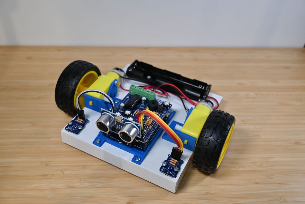 Front view of a white robot powered by 4AA batteries and propelled by two common yellow gear motors. The robot is controlled by an Arduino UNO R4 Minima with an attached ARPS-2 shield. The two optical sensor modules have been broken away from the ARPS-2 and mounted at the front corners of the robot chassis, and a SONAR module is plugged into the top of the ARPS-2.