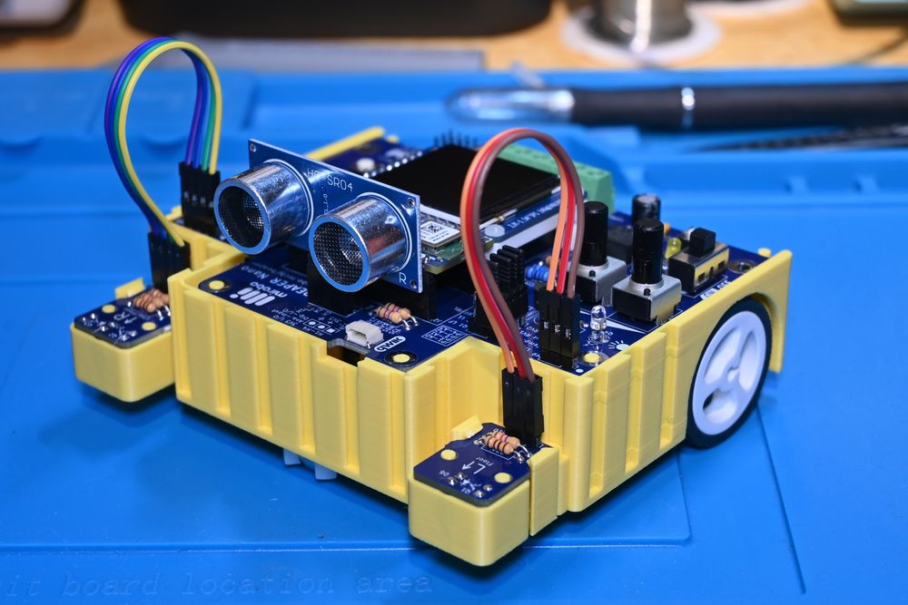 A yellow, 3D-printed BEAPER Bot robot sitting on a blue silicone mat. BEAPER Bot has dove-tail slots that allow sensors and servos to be mounted around its periphery. Two optical floor sensor modules are attached to the front of the robot and connected using colourful extension cables. An ultrasonic SONAR distance sensor module is mounted to a blue circuit board, centred between the floor sensor modules. The robot can be controlled by either a Raspberry Pi Pico mounted in BEAPER Pico circuit, or by an Arduino Nano ESP32 mounted in a BEAPER Nano circuit. This robot is built with a BEAPER Nano, and the Arduino Nano ESP32 is visible between the SONAR module and an LCD panel.
