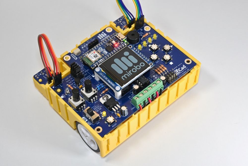 Rear-left view of a yellow 3D-printed BEAPER Bot robot controlled by an Arduino Nano ESP32 microcontroller mounted in a blue BEAPER Nano printed circuit board. The LCD display mounted between the Arduino and a green screw terminal strip is displaying the mirobo.tech logo.