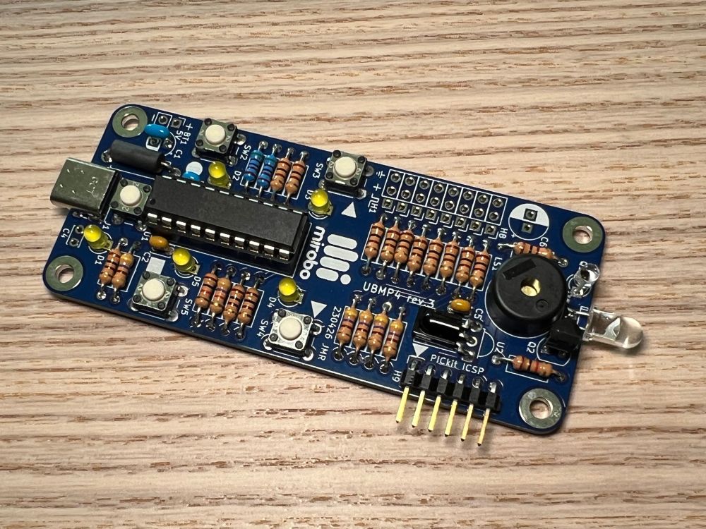 A fully assembled UBMP4 circuit sitting on a wooden tabletop. From left to right, the circuit features a USB type-C connector, a PIC16F1459 microcontroller surrounded by 5 pushbuttons and 5 LEDs, an expansion header area, an IR demodulator, an in-circuit programming header, a piezo speaker, an ambient light sensor, and an IR LED. 