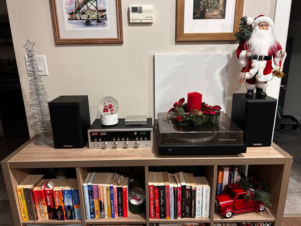 A bookshelf with a record player and some Christmas decorations- a silver wire tree, 2 snow gloves, a wreath with a candle, a Santa figure and a red truck with a tree