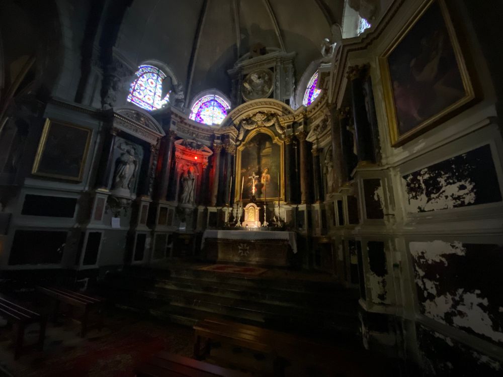 The interior of l’eglise Saint-Denis in Amboise, France. Dark walls are set with alcoves lined with statues or hung with paintings. The only illumination comes from stained-glass windows set up high, a single white light shining wide across an altar, and a blood-red point light source partly illuminating one alcove and statue. That statue is Saint Denis holding his head in his hands, as he was said to have survived being beheaded in Paris and to have carried his head to Amboise to be buried there.