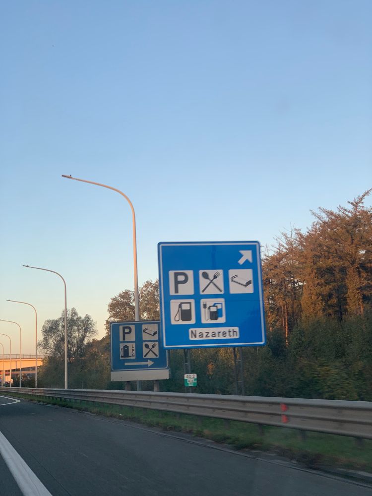 An exit sign on a freeway marks a rest stop called “Nazareth.” Internationally recognized symbols indicate the presence of parking, food, a place to sleep, and fuel. 
