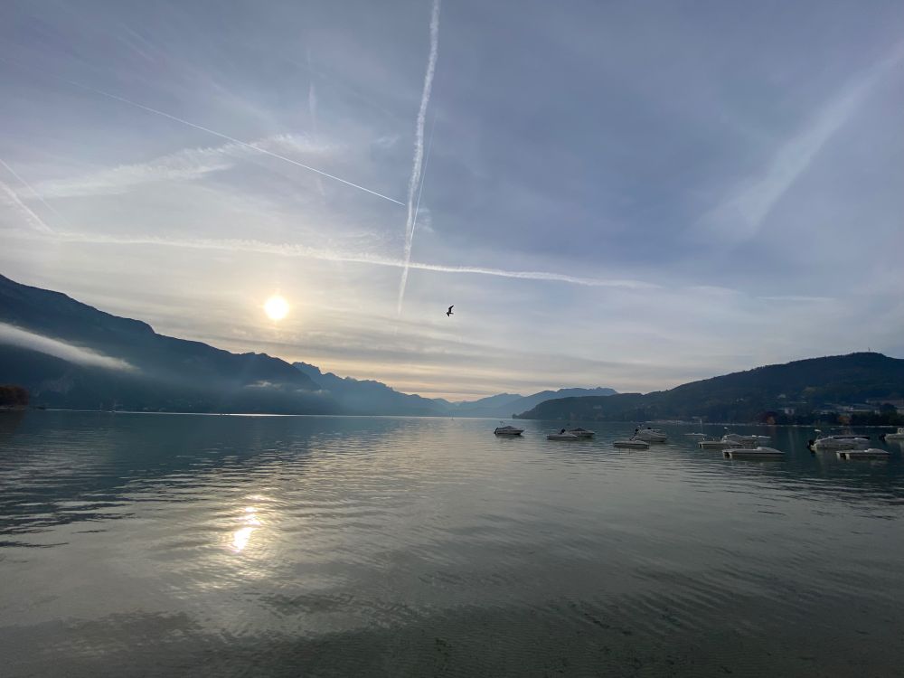 The broad vista of a lake spreads beneath blue morning sky streaked with cloud and jet contrails. To the right, pleasure boats are anchored out in the water. To the left, the sun hangs just above the distant peaks of the alps reflected in the rippling water.